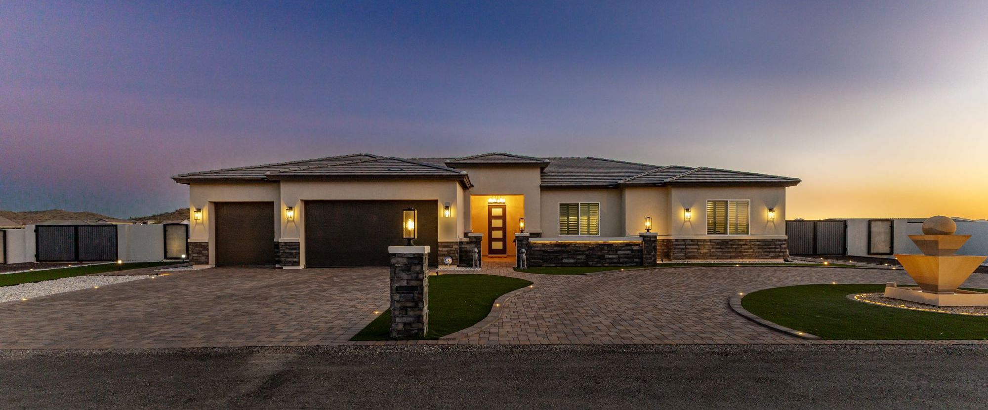 Single-story Arizona home glowing at twilight with a paver driveway and desert hills behind it